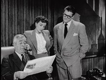 Clark Kent and Lois Lane look over Perry White's shoulder as he reads the paper